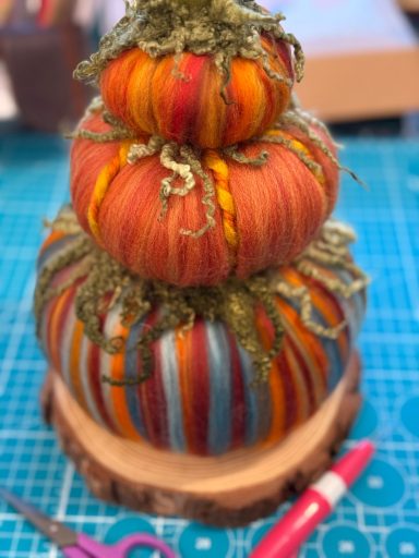 Three decorative pumpkins in vibrant colours, topped with green moss, on a wooden base.