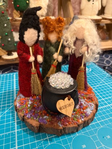 Three crafted figures in robes with brooms gather around a cauldron on a wooden base.