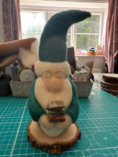 A handmade gnome figurine holding a stack of books, with a teal hat and glasses, on a wooden base.