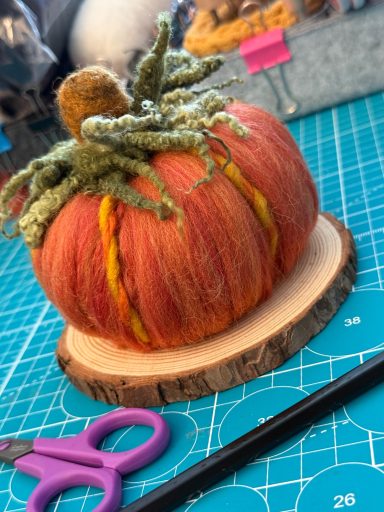 Decorative pumpkin with green moss and a wooden base, beside scissors on a blue background.