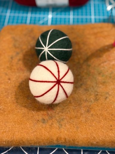 Two decorative felt balls, one green with white stitching and one cream with red stitching, on a mat.