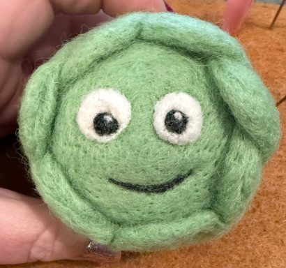 A green, round felted character with a smiling face and large eyes.