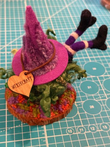A whimsical witch figurine with a purple hat and a heart tag, lying upside down.